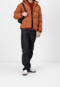 Hnědá prošívaná bunda s kapucí a zapínáním na zip, nošená přes červenou košili, doplněná tmavými džínami a bílými teniskami se šedými detaily.