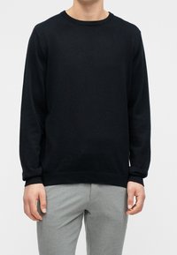 Personne portant un pull noir à manches longues et un pantalon gris clair, debout devant un fond clair uni.