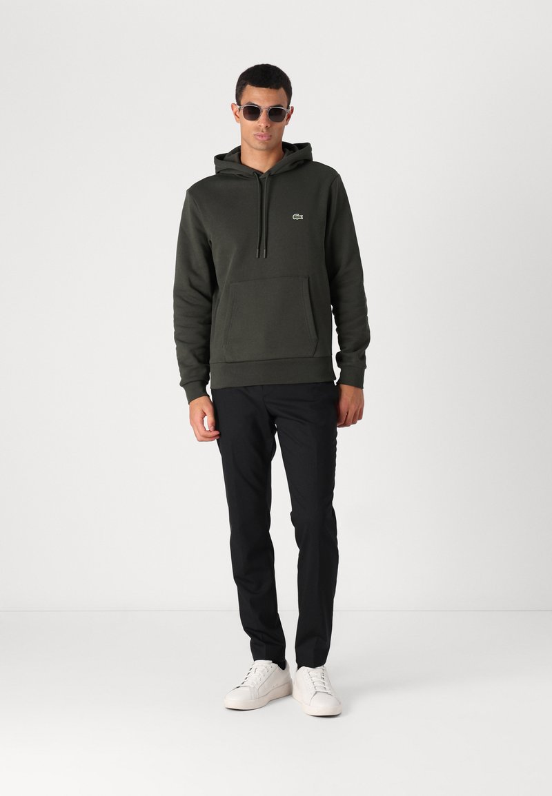 Sweat à capuche vert foncé avec une poche kangourou et un petit logo, associé à un pantalon noir et des baskets blanches. Modèle portant des lunettes de soleil.