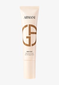 Beigefärgad hudvårdstub med ett vitt lock. Har svart text "ARMANI" och en stor brun logotyp. Texten anger "SKIN TINT långvarig & vård."
