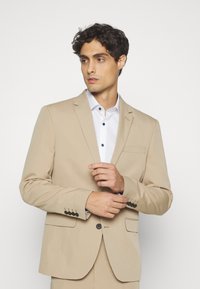 Beige színű zakó sima textúrával, egy gombos záródással és két oldalsó zsebekkel; fehér inggel párosítva, amely sötét gombokkal rendelkezik.