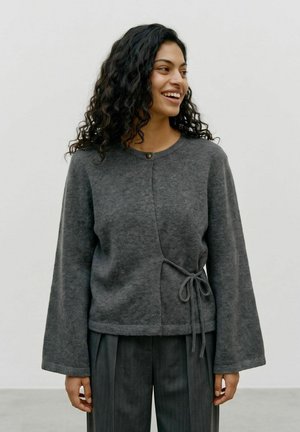 Femme aux cheveux bouclés foncés souriante, portant un pull cache-cœur gris foncé à manches longues et un pantalon ample gris foncé, debout devant un fond uni.