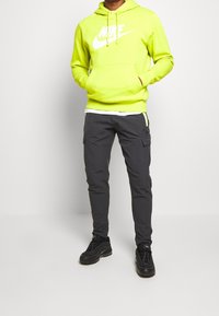 Person klädd i en neongul Nike-huvtröja, mörkgrå cargobyxor och svarta Nike-sneakers som står mot en vit bakgrund.