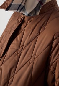 Veste matelassée marron avec couture en losanges et doublure à carreaux beige, noire et rouge visible à l'intérieur du col.