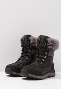 Czarne wodoodporne botki UGG z szarym futrem w środku, sznurowanym przodem i solidnymi podeszwami, zaprojektowane z myślą o zimnej pogodzie.