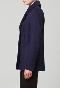 Tmavomodrý kabát z vlněné směsi s upraveným střihem, výstřihem s vykrojením, bočními kapsami a čistou siluetou. Texturovaná látka s hladkým povrchem.