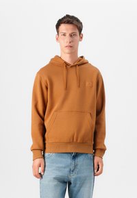 Hoodie marron en matériau doux, avec une poche avant, un capuchon à cordon de serrage et un petit patch logo sur la poitrine. Associé à un jean bleu clair.
