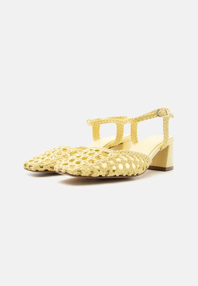Κίτρινα χαμηλά πέδιλα από πλεκτό δέρμα με λουράκια στον αστράγαλο και ασημένια αγκράφα σε απλό λευκό φόντο.