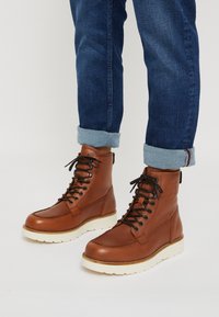 Brown leather lace-up boots with a white rubber sole, rounded toe, and black laces. Jeans with rolled cuffs showcase the boot design.