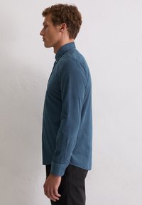 Türkisfarbenes Langarmshirt mit knöpfbarem Kragen, glatter Textur und seitlichen Schlitzen, kombiniert mit dunkler Hose.