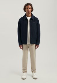 Navy fleece jack met rits, kraag, zijzakken en geborduurd logo; gedragen over een wit shirt en beige broek.