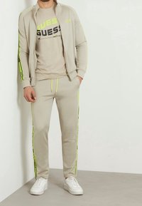 Tuta beige composto da una giacca con zip e pantaloni con cintura elastica. Accenti gialli brillanti lungo le maniche e i pantaloni. Sneakers bianche completano il look.