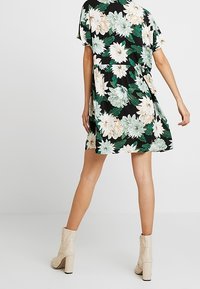 Blumenkleid mit schwarzem Hintergrund, das große weiße und grüne Blumen zeigt. Kurzärmlig, lockere Passform und knielanges Design. Beigefarbene High-Heels.