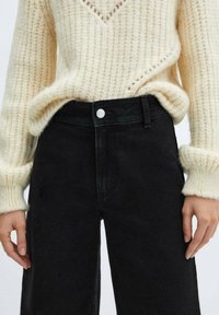 Persona vistiendo un suéter de punto color crema metido en unos jeans negros de cintura alta, con botones visibles y trabillas para el cinturón, con las manos relajadas a los lados.