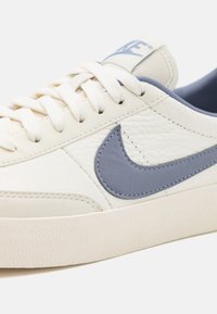 Bijele kožne tenisice s svijetloplavim swoosh logotipom, teksturiranim gornjim dijelom, ravnim vezicama i gumenenim potplatom. Minimalistički dizajn s kontrastnim akcentima.