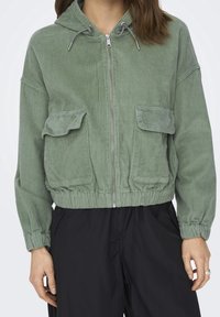 Veste en velours côtelé vert avec fermeture éclair à l'avant, capuche à cordon de serrage et deux poches à rabat à l'avant. Poignets et ourlet élastiques. Texture douce et coupe décontractée.