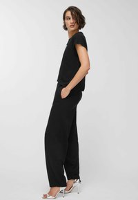 Kvinde stående sidelæns iført sort kortærmet top, sorte bukser med hænderne i lommerne og sølvfarvede højhælede sandaler på en ensfarvet baggrund.