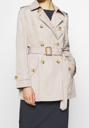 Beiger zweireihiger Trenchcoat mit großen Knöpfen und Gürtel, getragen über einem schwarzen Kleid mit weißen Punkten, Hand in der Tasche.
