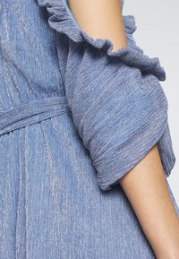 Robe en tissu texturé bleu scintillant avec manches bouffantes et bord volanté sur un bras visible sur fond uni.