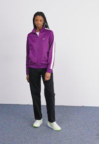 Jachetă violet cu fermoar, cu dungi albe, asortată cu pantaloni negri și adidași albi. Prezintă un guler înalt și logo-ul Nike pe piept.