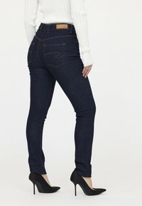 Dunkelblaue Jeans mit schmaler Passform, hohe Taille, Gesäßtaschen mit Stickdetails, kombiniert mit schwarzen High Heels.