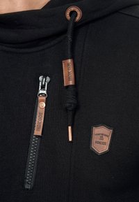 Sudadera negra fabricada en un tejido suave, con un bolsillo con cremallera prominente, detalles de cuero y un parche con el logo de la marca en el pecho.