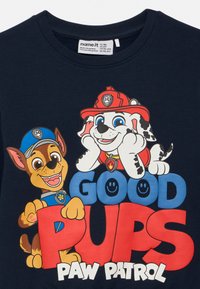 Maglietta blu navy con cani dei cartoni animati colorati. Il testo recita "BUONI CUCCIOLI" in lettere grassetto rosse e blu, con un'icona di un viso sorridente e "PAW PATROL" sotto.