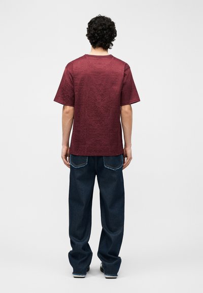 Ανδρας με σγουρά μαλλιά που φοράει ένα υφασμάτινο βυσσινί κοντομάνικο πουκάμισο και άνετο σκούρο μπλε τζιν, όρθιος με την πλάτη προς τα εμάς σε λευκό φόντο.