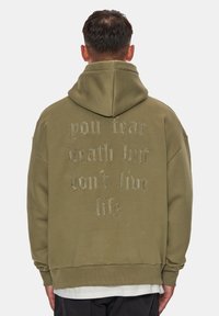 Felpa con cappuccio verde oliva realizzata in tessuto morbido, con cappuccio ampio e grande scritta ricamata sulla schiena. Orlo arrotondato e vestibilità comoda.