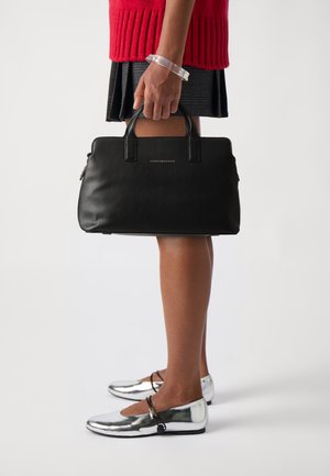 Femme en pull rouge et jupe noire tenant un sac à main noir Tommy Hilfiger, portant des ballerines argentées de style Mary Jane et un bracelet transparent.