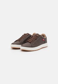 Brune lærersneakers med såle i hvitt gummi, med brune snørebånd og en rød logoak accent. Glatt tekstur med perforerte detaljer.
