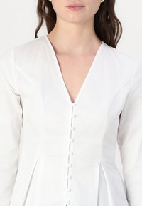 Balta suknelė su giliu V formos iškirpimu, mygtukų užsegimu ir ilgomis rankovėmis. Turtinga klostuota sijonas ir švelnus medvilnės audinys.