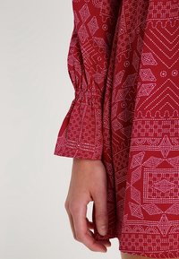 Rotes Kleid mit weißen geometrischen Mustern, das über eine geraffte Ärmelgestaltung verfügt. Der Stoff wirkt glatt und hat eine weiche Textur.