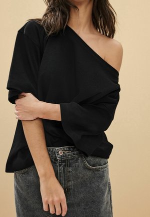Femme portant un haut noir ample épaules dénudées et une jupe en jean délavée taille haute, la main reposant sur le bras opposé.