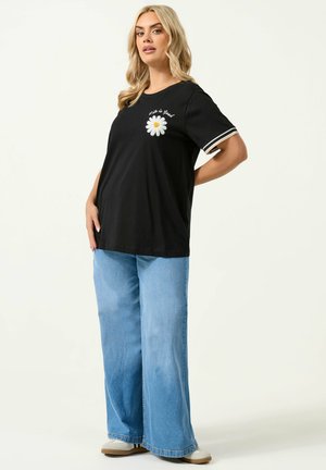 Kobieta o blond włosach ubrana w czarną koszulkę z grafiką stokrotki i napisem "Life is Good", jasnoniebieskie dżinsy z szerokimi nogawkami oraz białe trampki.