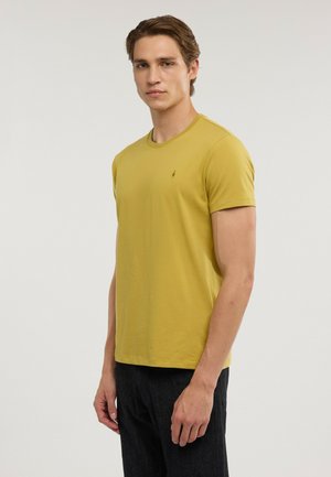 Joven con una camiseta amarilla mostaza lisa de manga corta y pantalones oscuros, de pie frente a un fondo claro y liso.