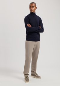 Maglione blu navy con zip in un design aderente, abbinato a pantaloni beige chiaro e sneakers marroni, che mostrano un look casual e moderno.