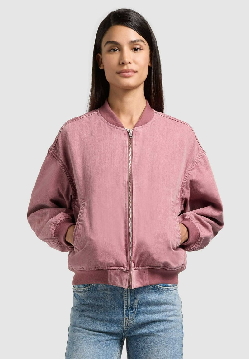 Jeune femme aux longs cheveux foncés portant une veste bomber rose zippée et un jean bleu clair, les mains dans les poches de la veste, fond neutre.