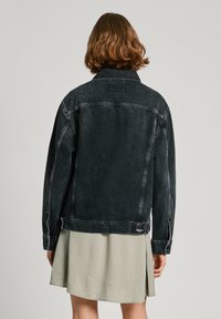 Svart denimjacka med en strukturerad design, långa ärmar, synlig sömnad och en etikett vid fållen; bärs över en ljusmönstrad klänning.