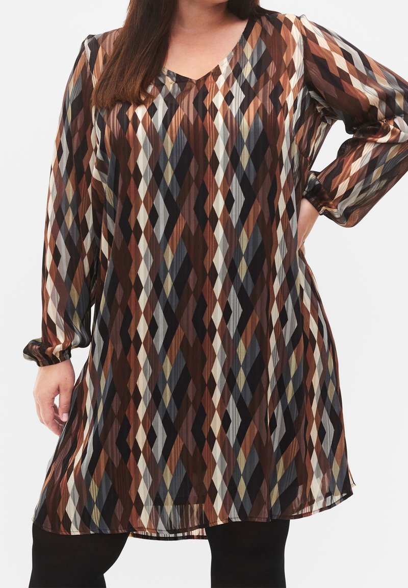 Femme portant une robe à manches longues, longueur genou, avec un motif géométrique en forme de losange aux tons marron, beige, noir et gris.