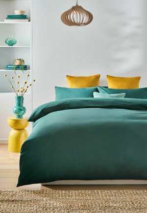 Türkisfarbene Bettwäsche mit senfgelben Kissen auf einem Bett, darüber ein hängendes Holzlicht, gelber Beistelltisch mit grüner Vase mit gelben Blumen.