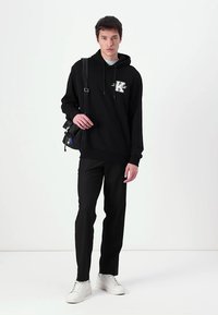 Hanorac negru cu logo grafic, asortat cu pantaloni negri și adidași albi. Modelul poartă o geantă mică neagră, stând în fața unui fundal simplu.