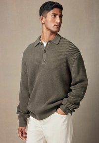 Olivengrøn, ribbet strikket pullover med krave og tre knapper. Lange ærmer med manchetter, parret med lyse bukser.