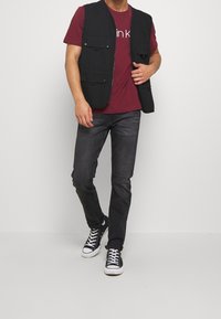 Czarna kamizelka z wieloma kieszeniami z przodu, noszona na bordowej koszuli, w połączeniu z ciemnymi dżinsami i czarno-białymi trampkami.