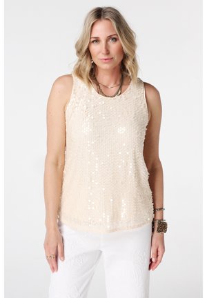 Blonde vrouw die een beige mouwloze top met pailletten draagt, witte geborduurde broek, gelaagde kettingen en armbanden tegen een effen achtergrond.
