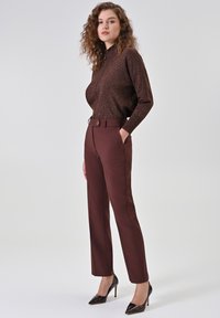 Maglione marrone scintillante con collo alto abbinato a pantaloni bordeaux su misura con dettaglio a bottone e tacchi neri a punta.