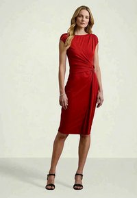 Frau mit langen blonden Haaren, die ein enges rotes knielanges Kleid mit seitlichem Knoten und schwarze High Heels trägt, steht vor einem schlichten Hintergrund.