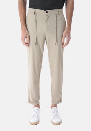 Uomo che indossa pantaloni beige con coulisse e orli risvoltati, e sneaker bianche, in piedi davanti a uno sfondo semplice.