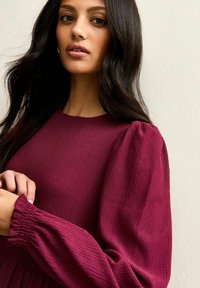 Burgundowa dzianinowa sukienka z teksturowanej tkaniny, z długimi bufkami oraz marszczonymi mankietami. Styl obejmuje okrągły dekolt i dopasowaną górę.