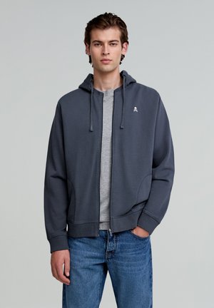 Grijze zip-up hoodie met een capuchon, voorzien van een klein doodskoplogo op de borst, gedragen over een grijs T-shirt, gecombineerd met een blauwe spijkerbroek.